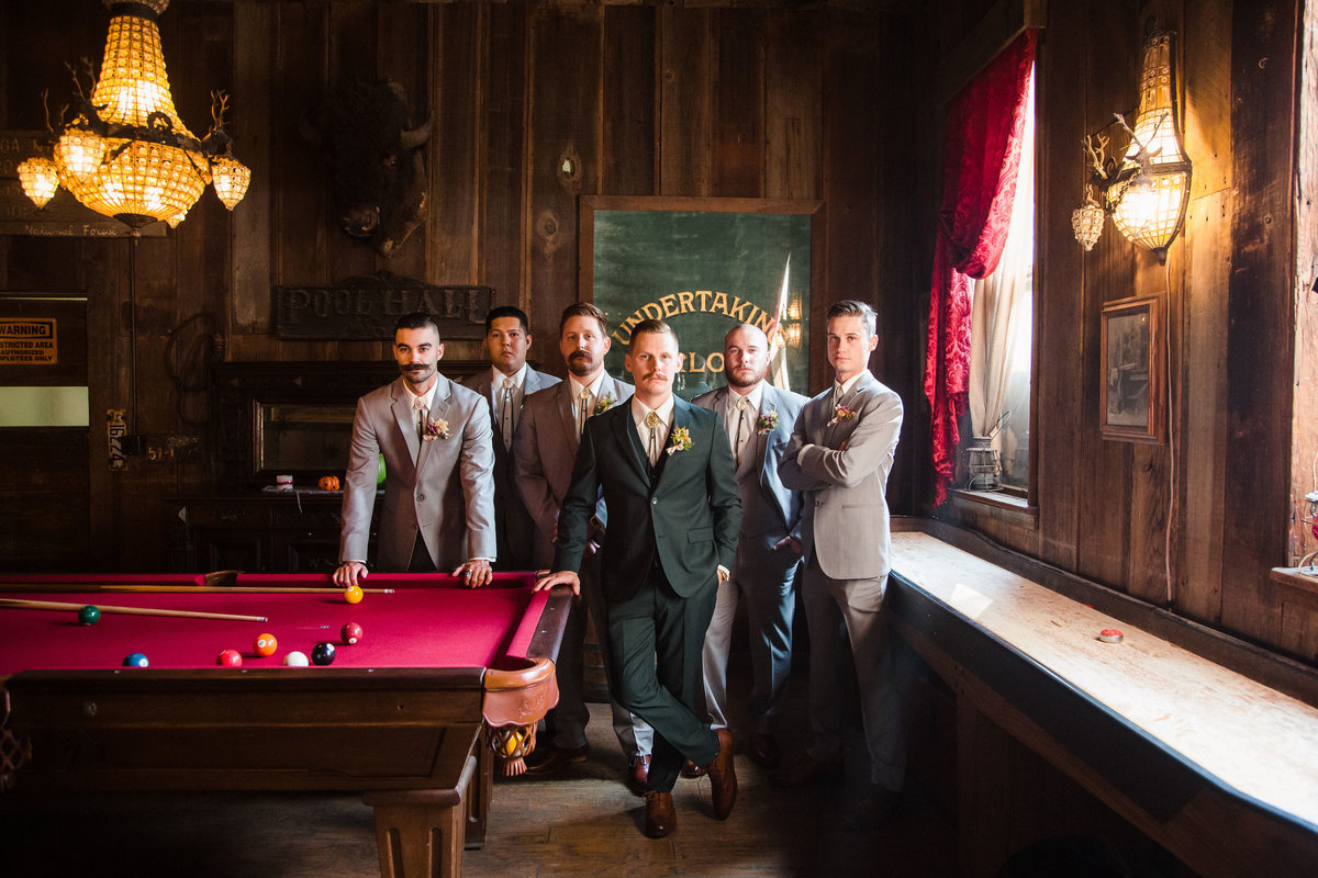 Mustached groom and his groomsmen standing at a pool table in a rustic pool hall,  in their wedding suits, before the ceremony