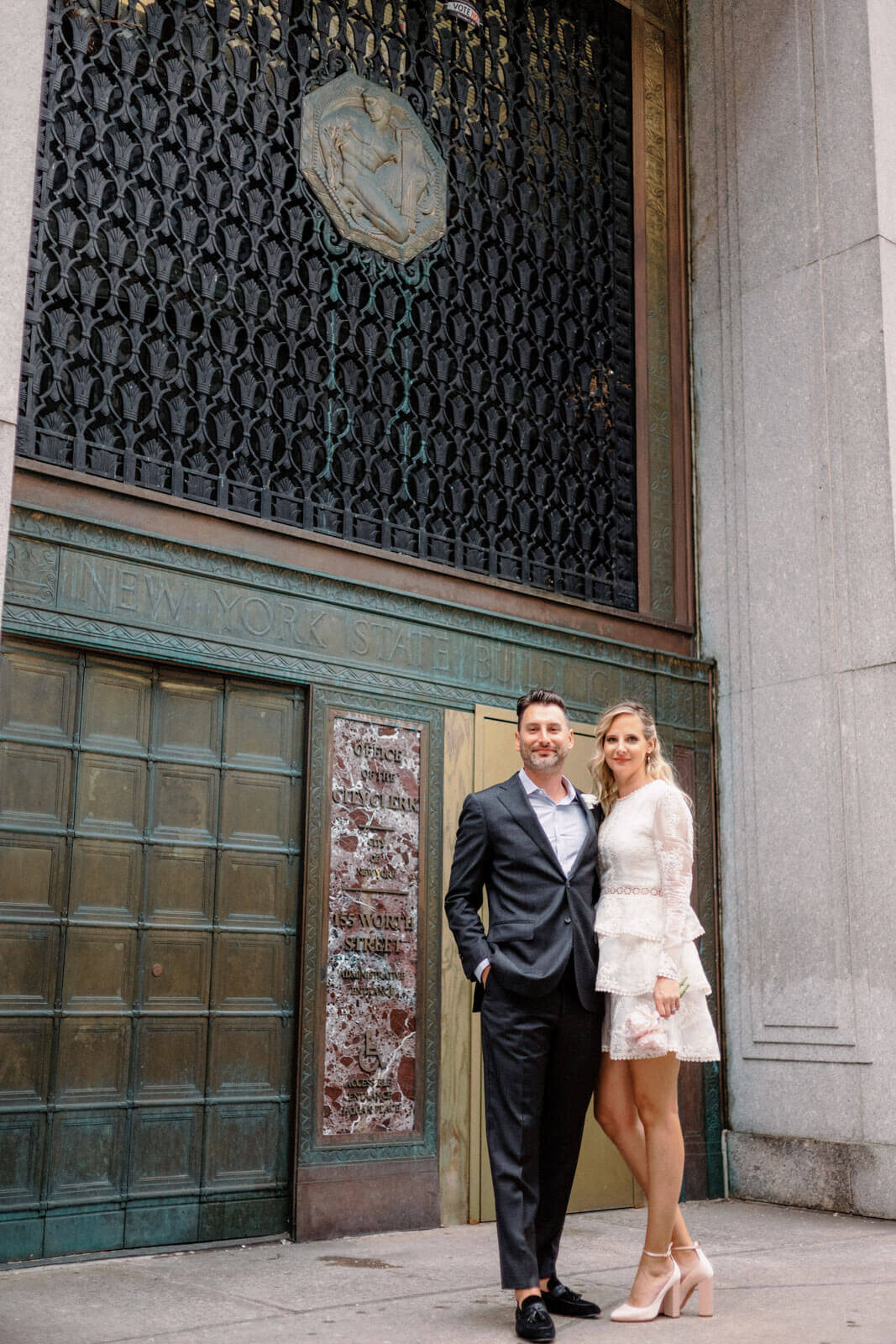 The bride and groom are standing close, smiling, outside the NY City Hall exit door after their elopement. Image by Jenny fu Studio