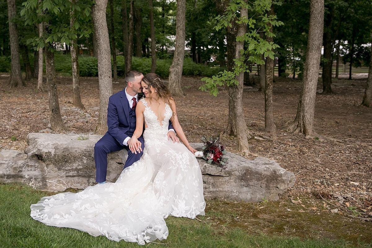 The groom, wearing a navy suit and burgundy tie sits behind the bride  on a limestone rock outcropping in a cedar glade wooded area at Saddle Woods Farm. The bride is wearing a long sleeveless lace wedding gown with a deep plunging neckline and is holding a bridal bouquet of burgundy and blush roses, red peonies and burgundy ranunculus wrapped in an ivory satin ribbon.