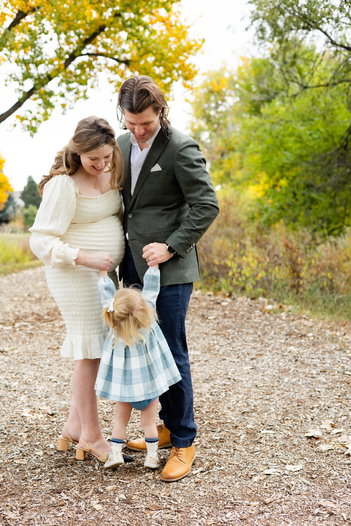 A mom and dad hold each other close and their toddler daughter stands in front of them reaching up to be picked up.
