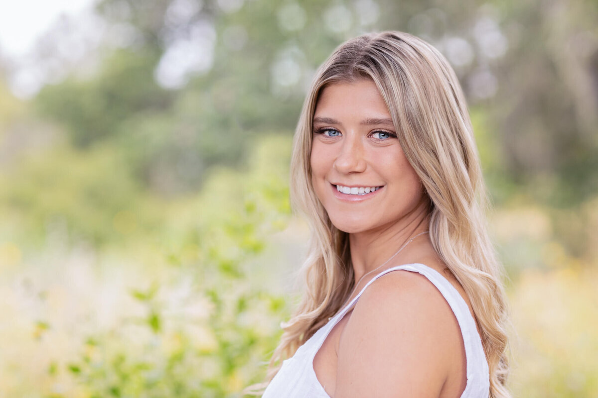 A woman with long blonde hair smiles while standing outdoors. She is wearing a white sleeveless top. The background is blurred with green foliage.