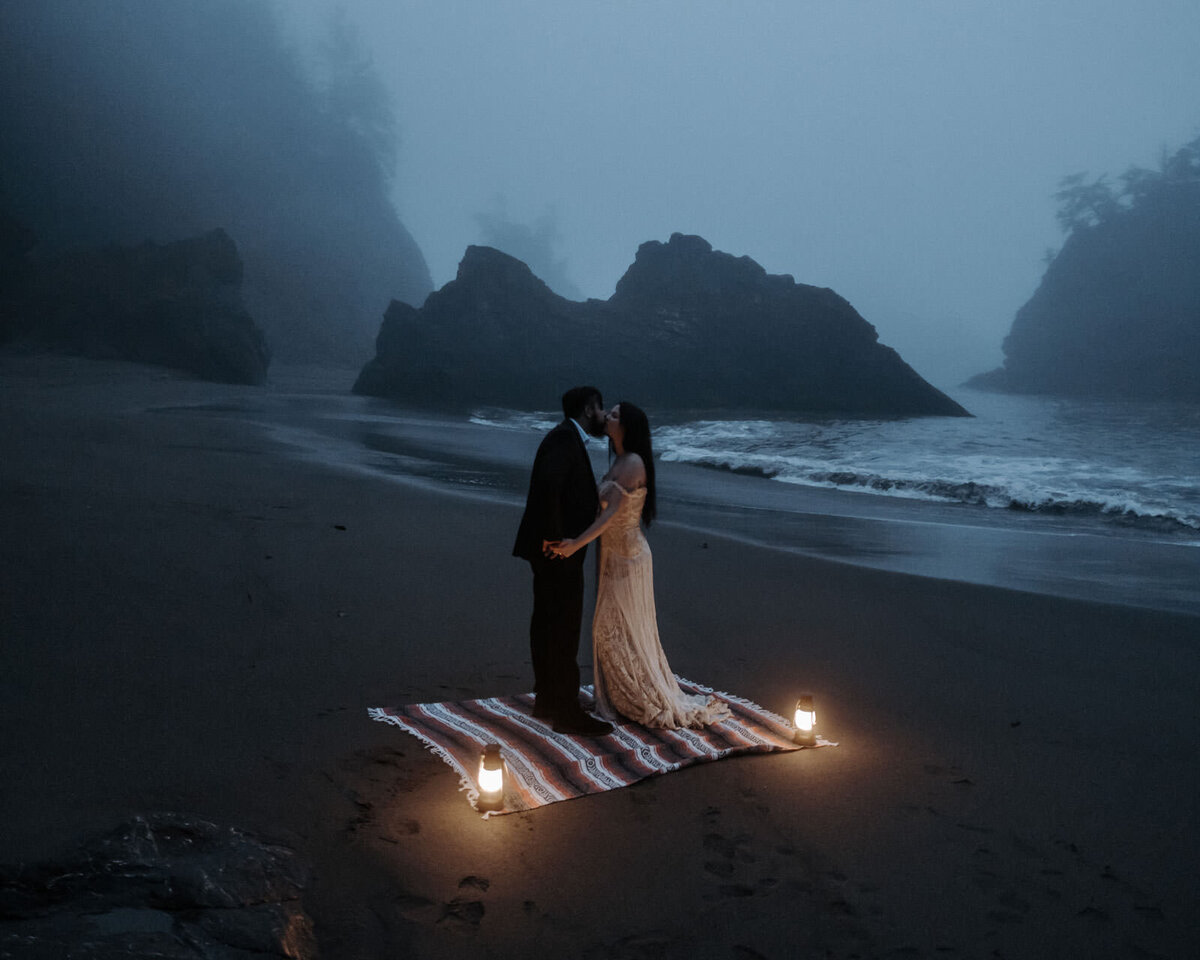 A bride and groom holding hands at Secret Beach in Oregon in the fog. It's dark and there are lanterns and a blanket on the ground.