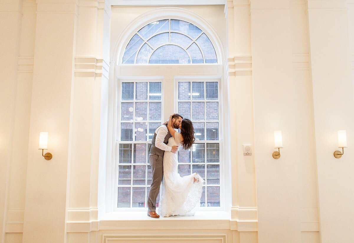 Standing on the deep window sill in front of a 14 foot arched window at Noelle Hotel in Nashville the groom wearing gray pants and a gray vest with a white shirt kisses the bride who is wearing a strapless mermaid gown with her foot kicked up in the air.