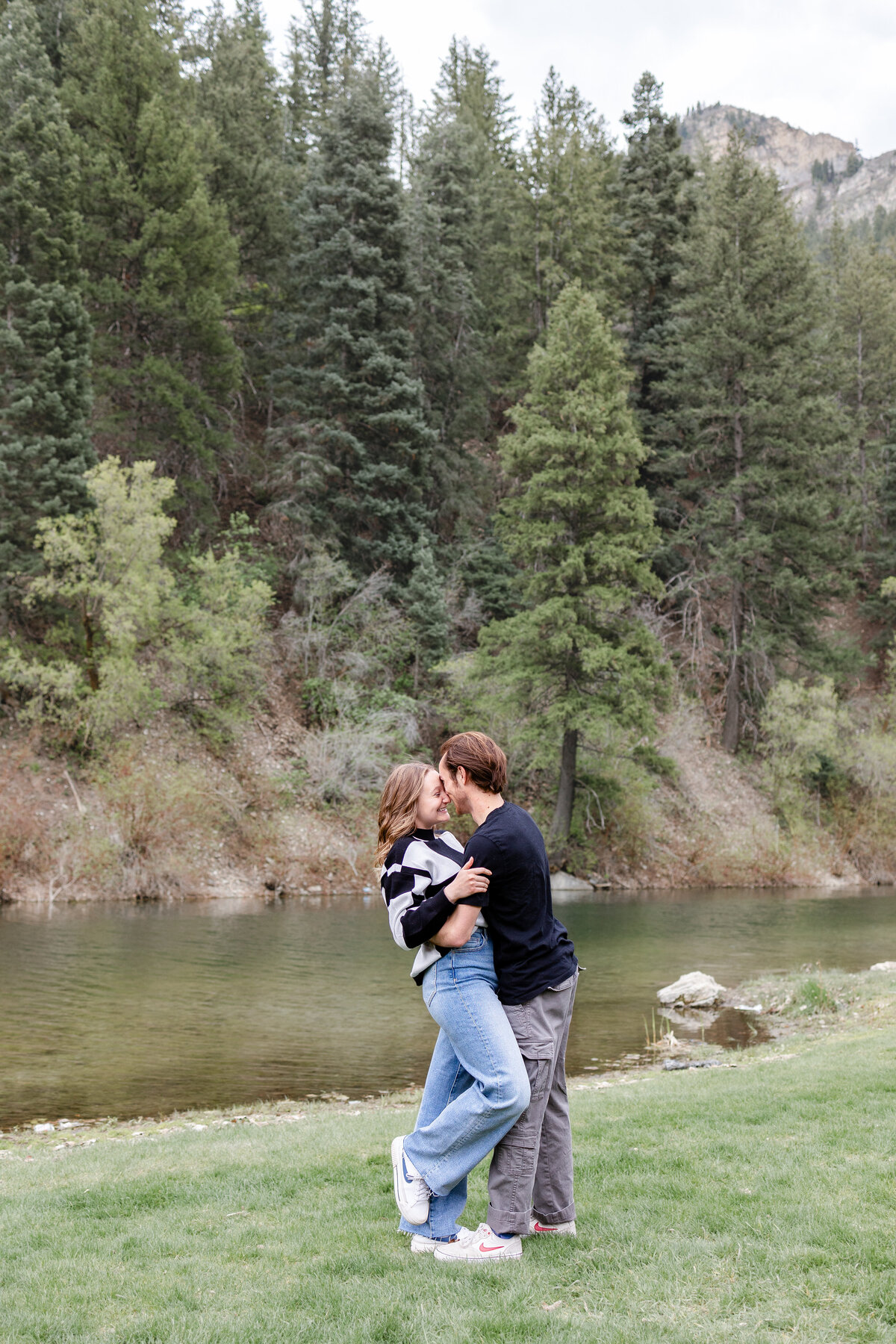 A couple sharing a romantic kiss beside the river at Vivian Park, framed by lush greenery and towering pine trees.