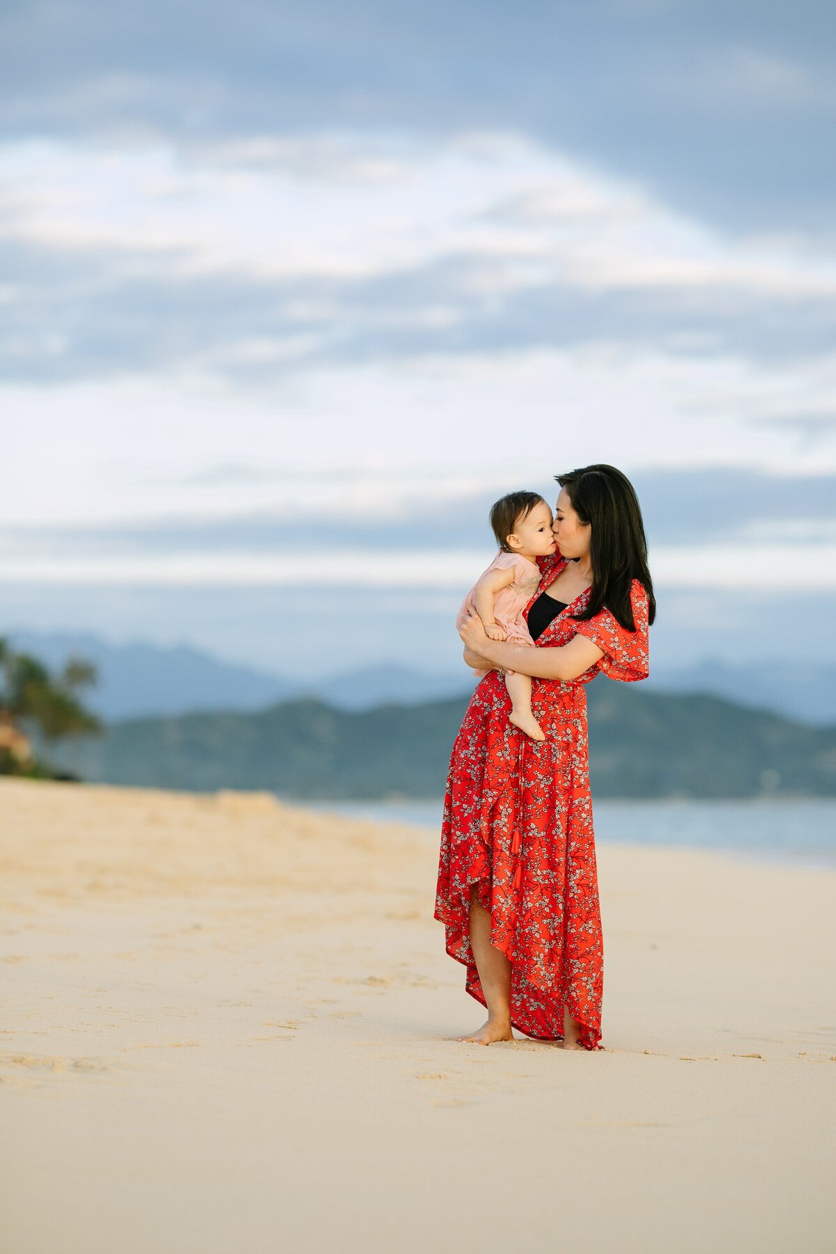 mom in a red dress kisses her baby daughter at lanikai beach