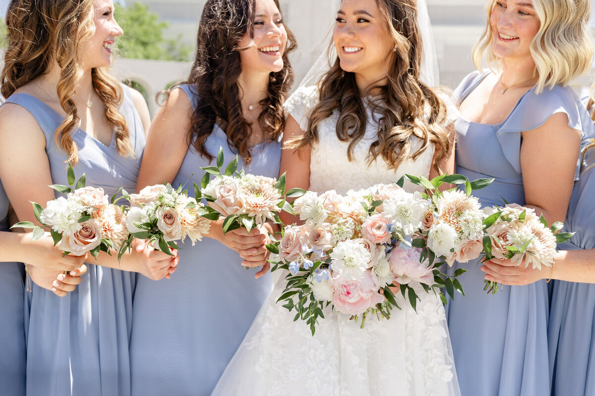The bride and her bridesmaids, dressed in soft blue gowns, hold delicate floral bouquets and smile brightly.