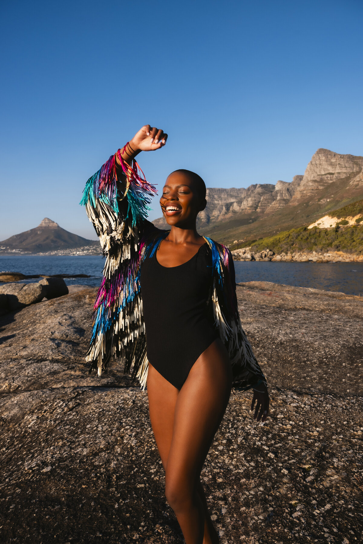 Smiling model enjoying golden hour on the rocks in Cape Town, wearing a colorful fringe jacket over a black bodysuit, with clear blue skies and mountain views in the background.