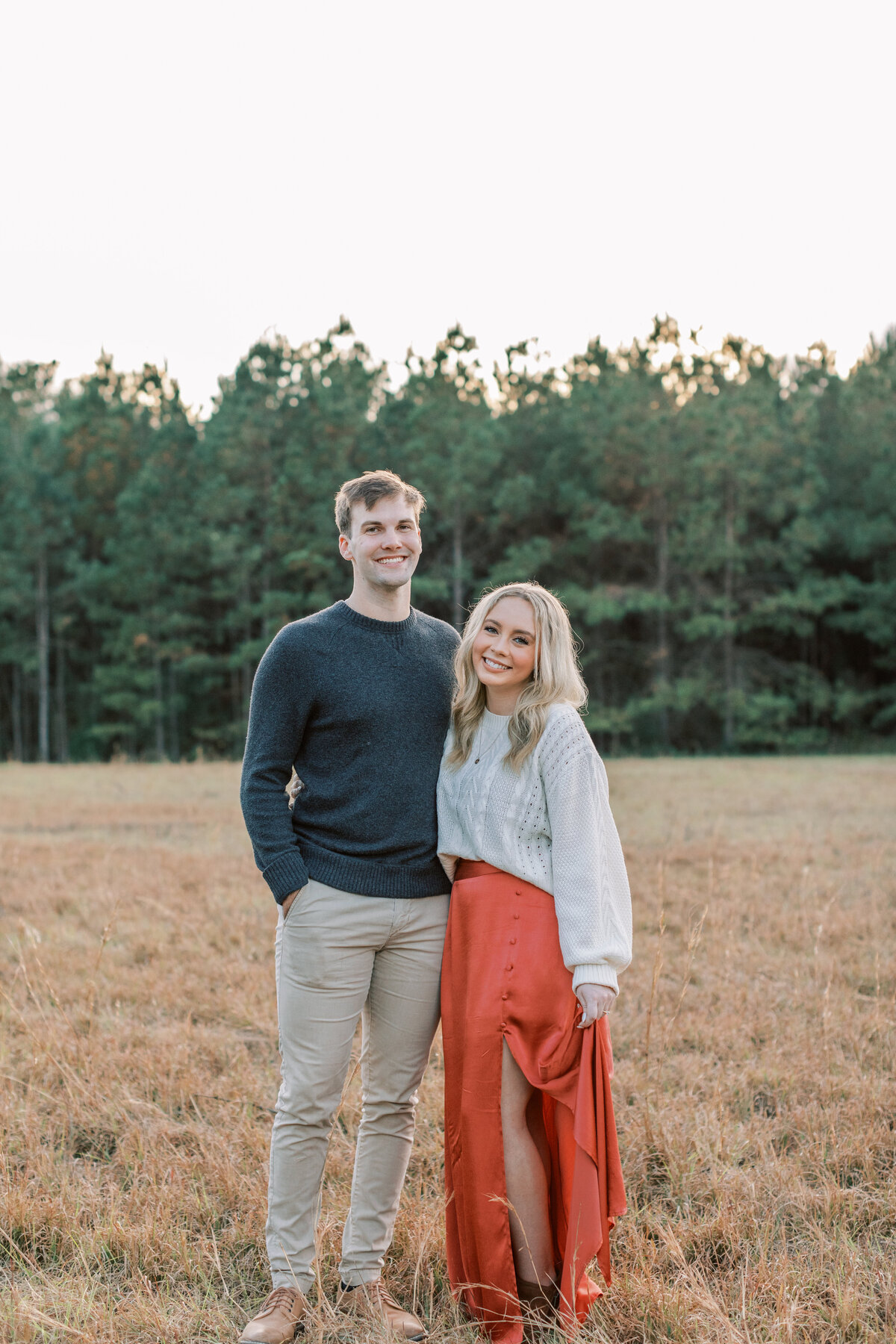A couple stands in a field while smiling at the camera.