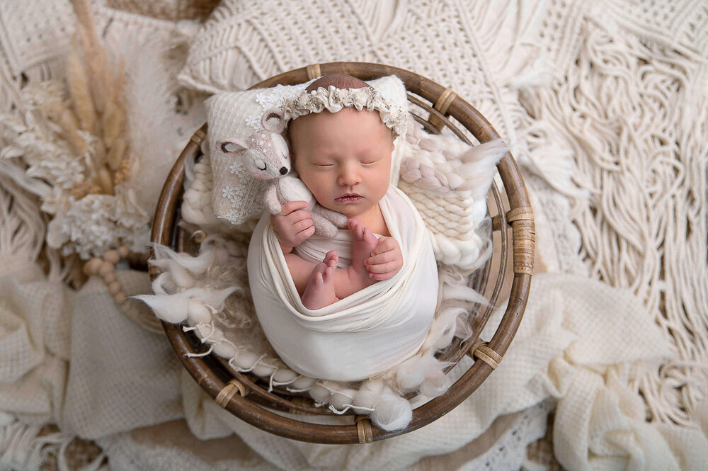 Baby girl wrapped in cream holding a little fawn in a boho-style set up for her newborn photography session.