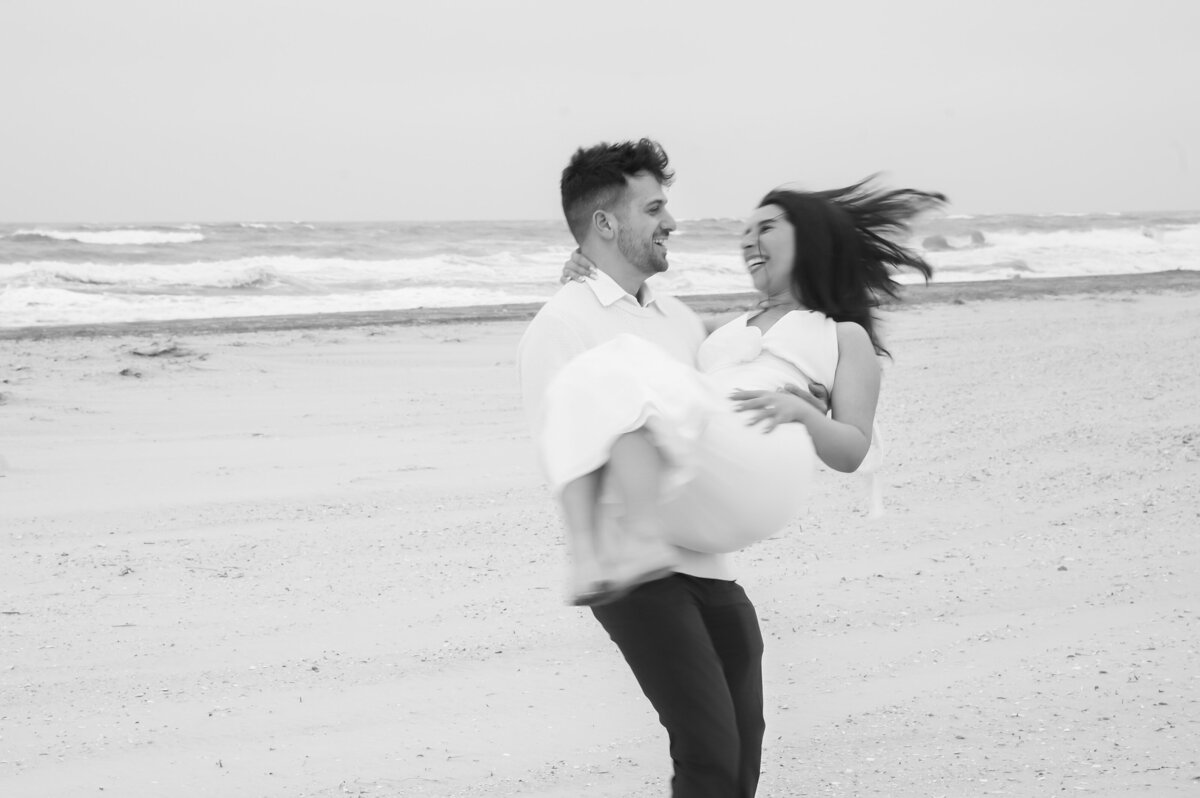 A man in a white shirt lifts a woman in a white dress while standing on a sandy beach with waves in the background.