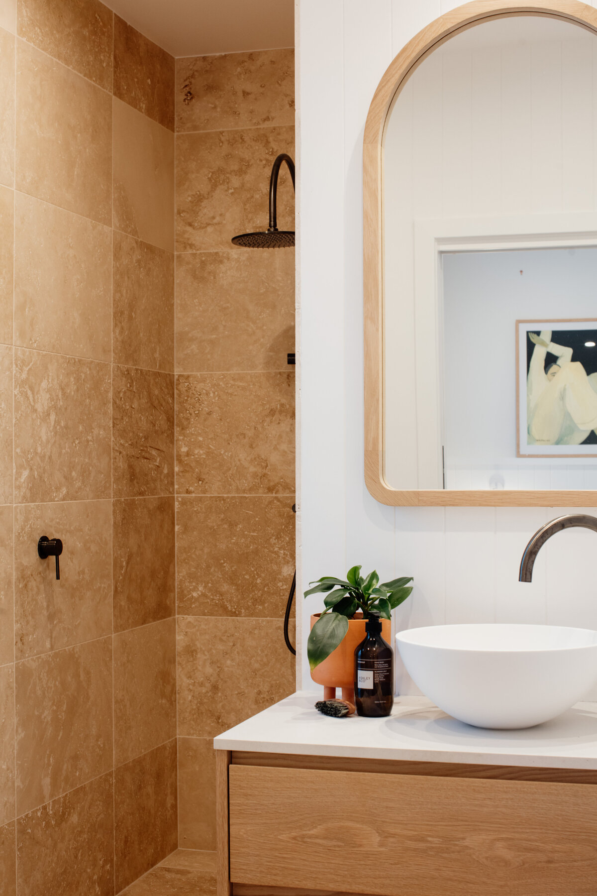 Modern bathroom featuring stone tiles, a wooden vanity, round mirror, white sink, and a potted plant—a testament to bespoke interior design in Newcastle NSW.