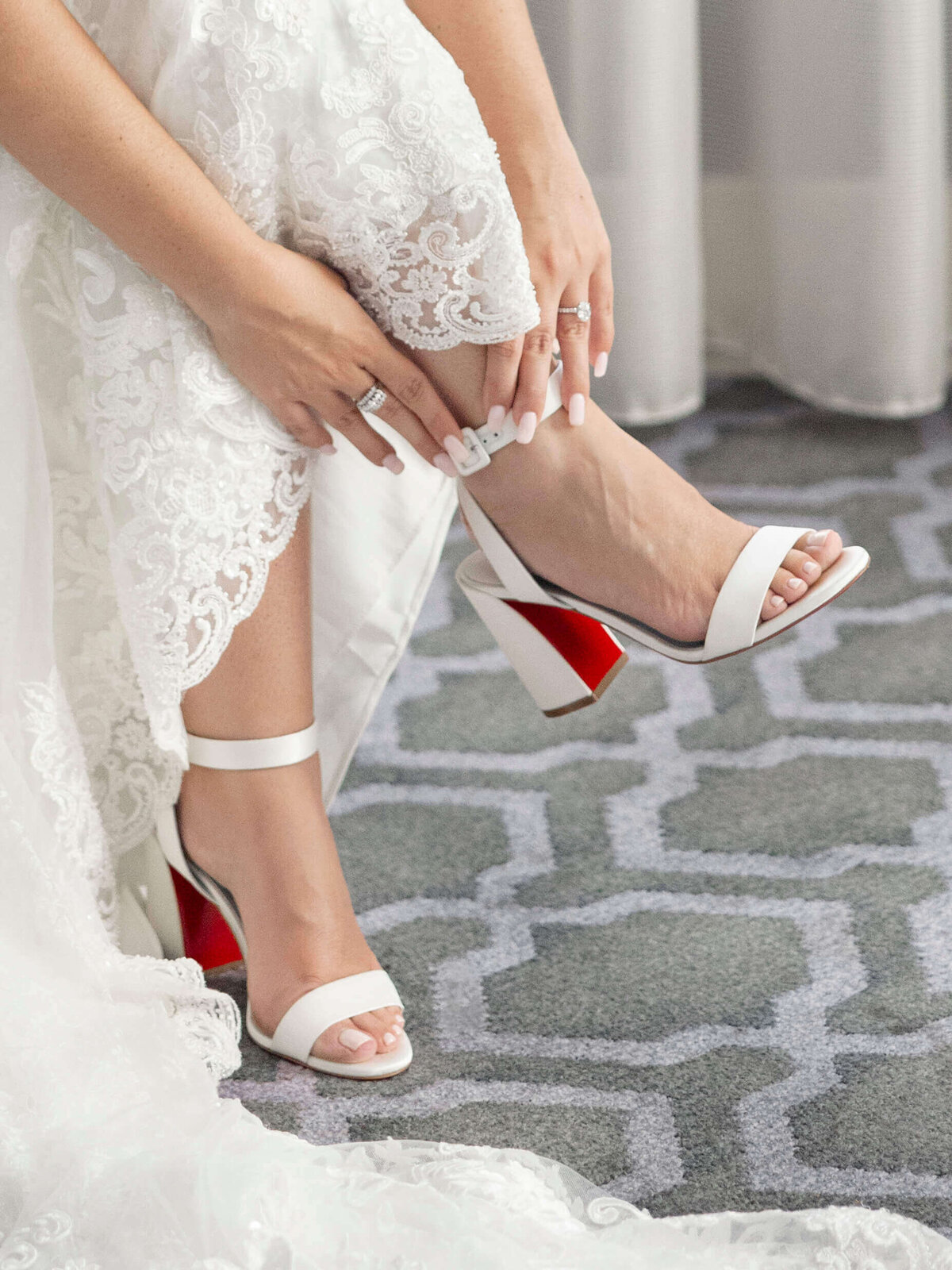 A bride in a lace wedding dress adjusts her white block-heeled sandals with red soles. Her nails are manicured with a light polish, and she is sitting on a geometric-patterned carpet.