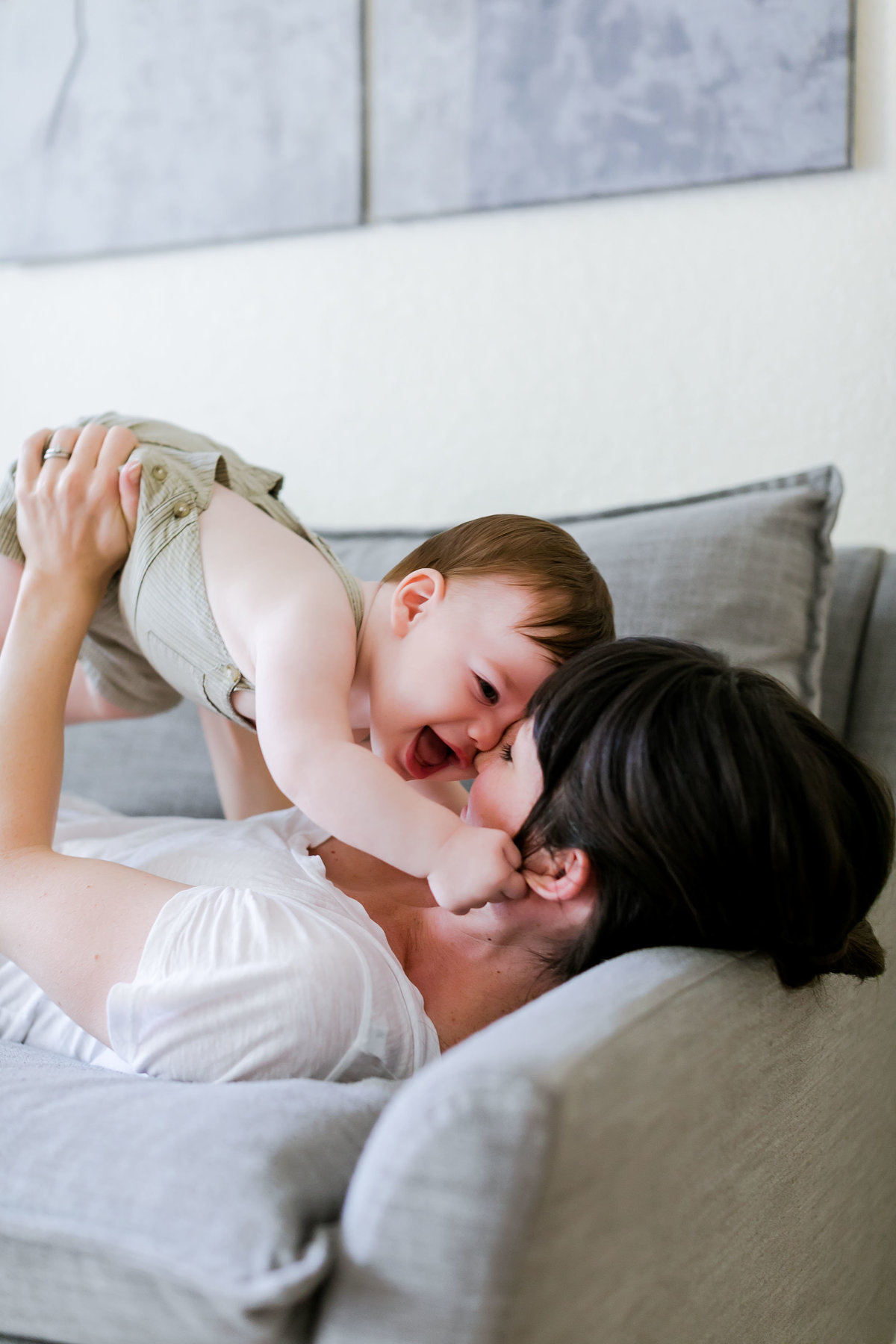 A loving mother in soft white cradles her baby boy in sage green, smiling as they play together on the couch during an in-home lifestyle session with Brooke Tucker photography  