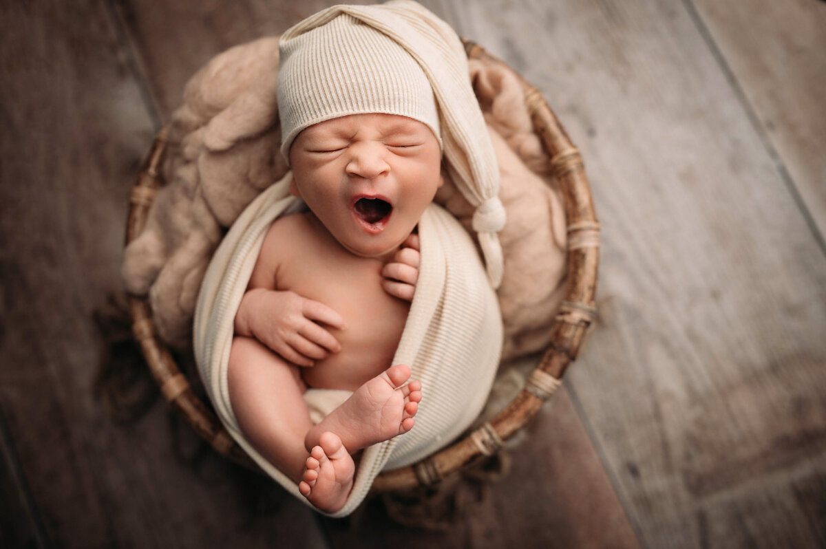 Fairfield CT newborn photographer captures sleeping and yawning baby wearing a beige cap and wrapped in matching fabric inside of a  brown bowl prop
