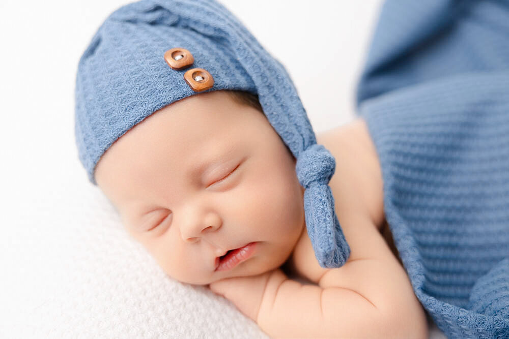 Baby newborn boy sleeping on belly on a white waffle blanket  and wearing a blue hat. 