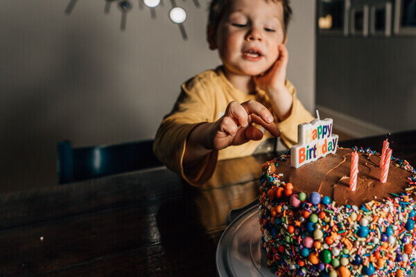 A three year old boy reaching to grab a large, colorful sprinkle off his birthday cake.