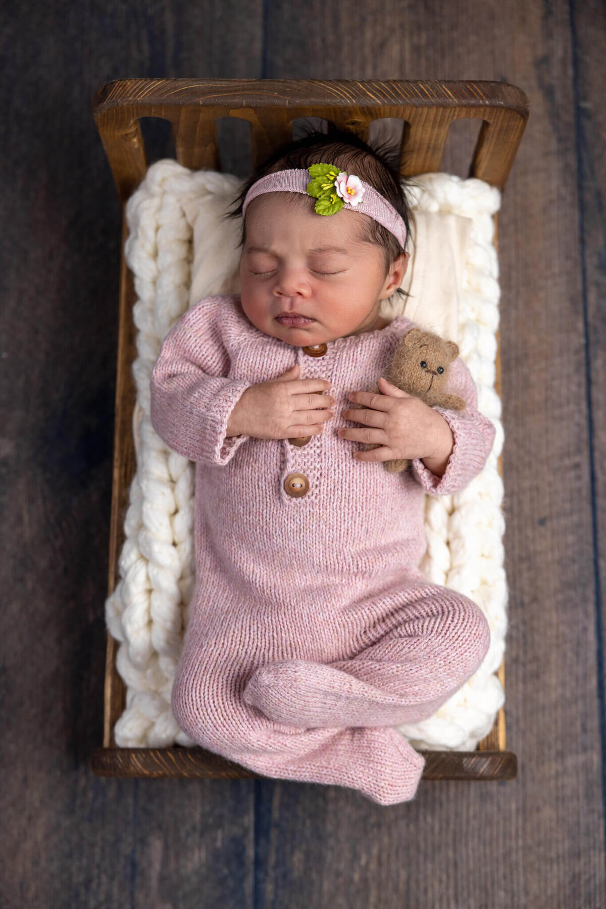 newborn baby with dark hair in a knitted pink romper asleep on a tiny wooden bed holding a tiny teddy bear