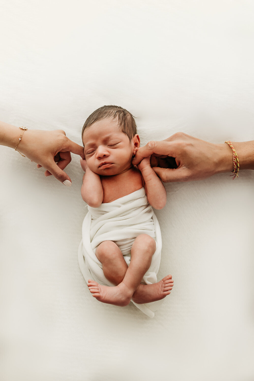 newborn baby wrapped in white swaddle. a parents hand holds each of hers in an overhead photo.