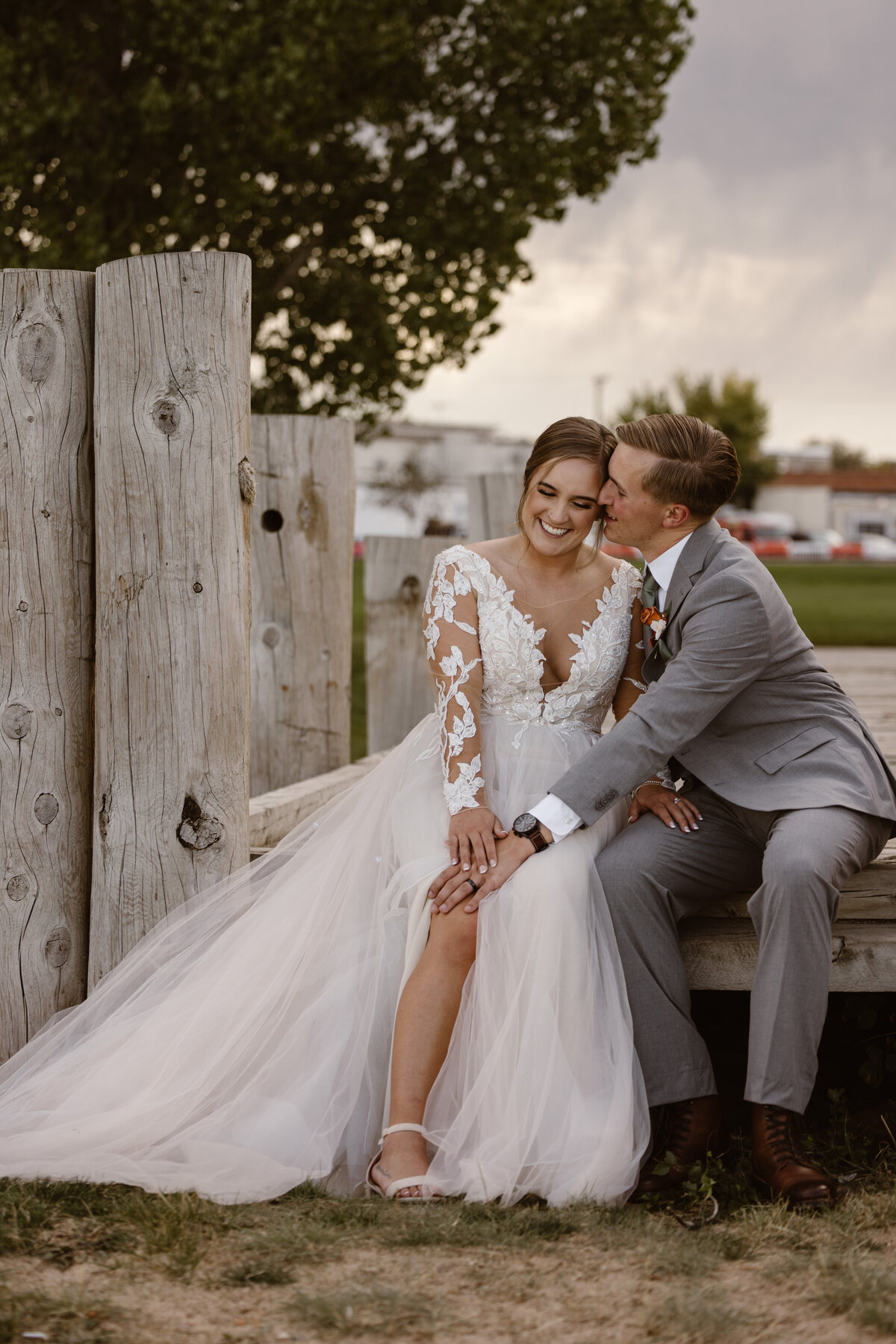 Groom kisses bride on the cheek as she smiles, sitting together on rustic wood beams during golden hour at their Colorado wedding.