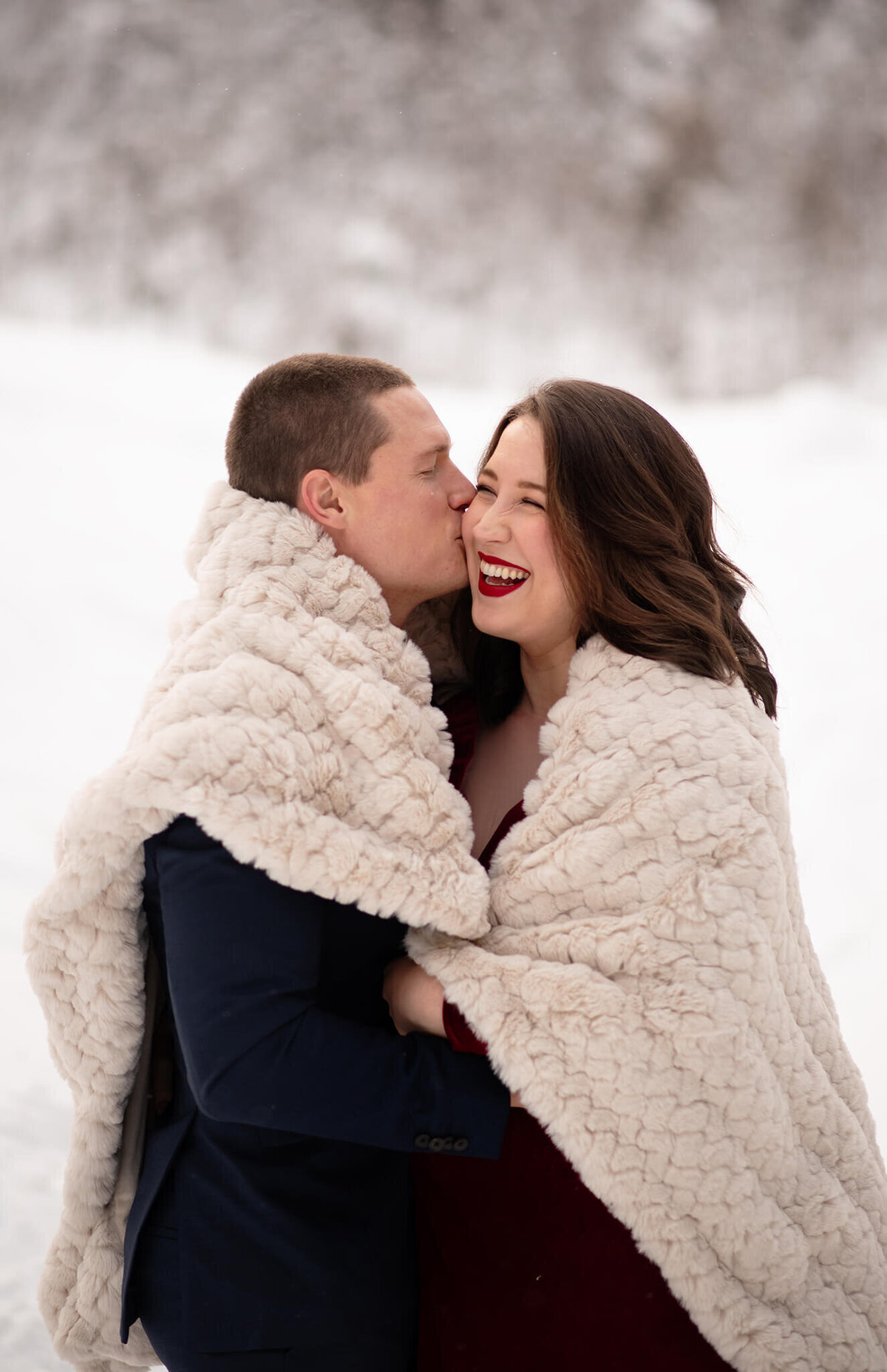 A husband and wife snuggle under a blanket, their faces are close together. The husband kissed her cheek and she laughs. There is a snowy backdrop in Sun Peaks. Taken by Brittany Danielle Photography