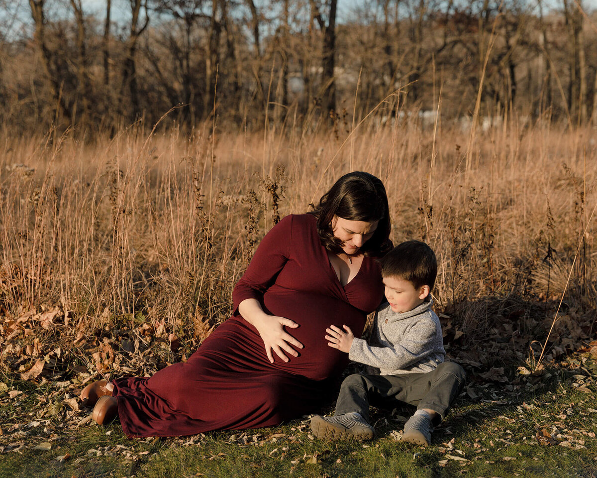 Pregnant Mom in maroon dress sitting on the grass as her toddler sons touches her baby belly.