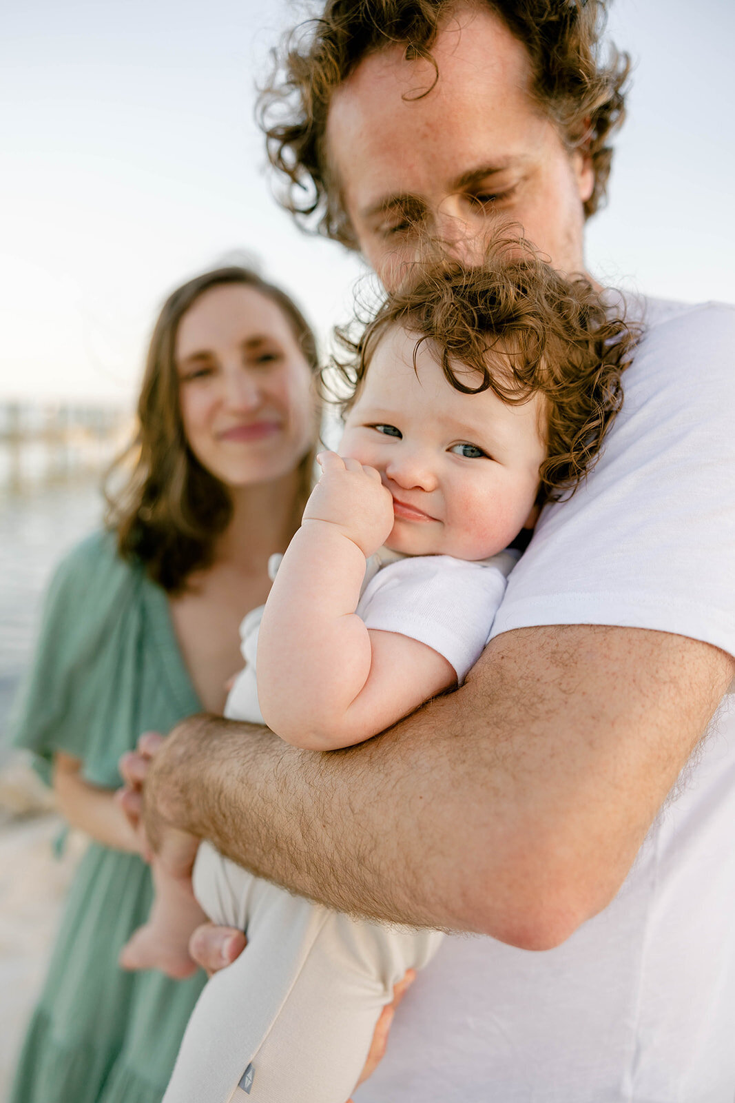 A father kisses his curly-haired baby while the mother smiles in the background during a Florida Keys family photography session.