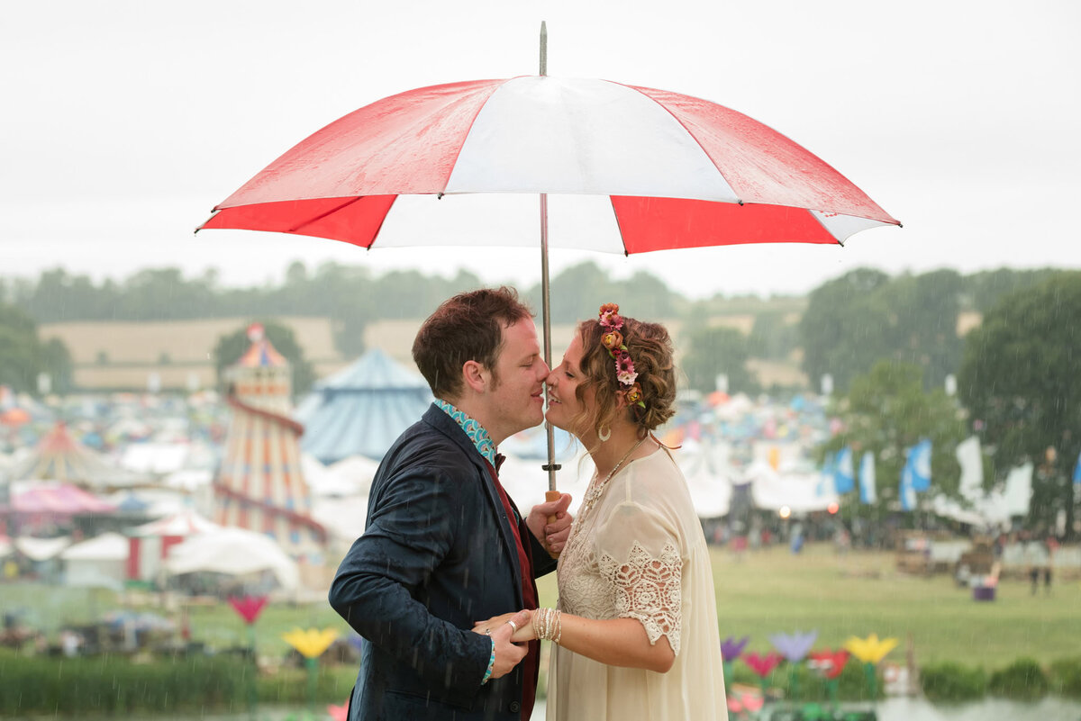Bride and groom in boho outfits, shltering under an umbrella, with a festival view behind them