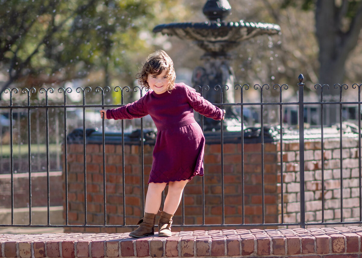 A child in a maroon dress and boots standing by a metal fence with a fountain in the background.