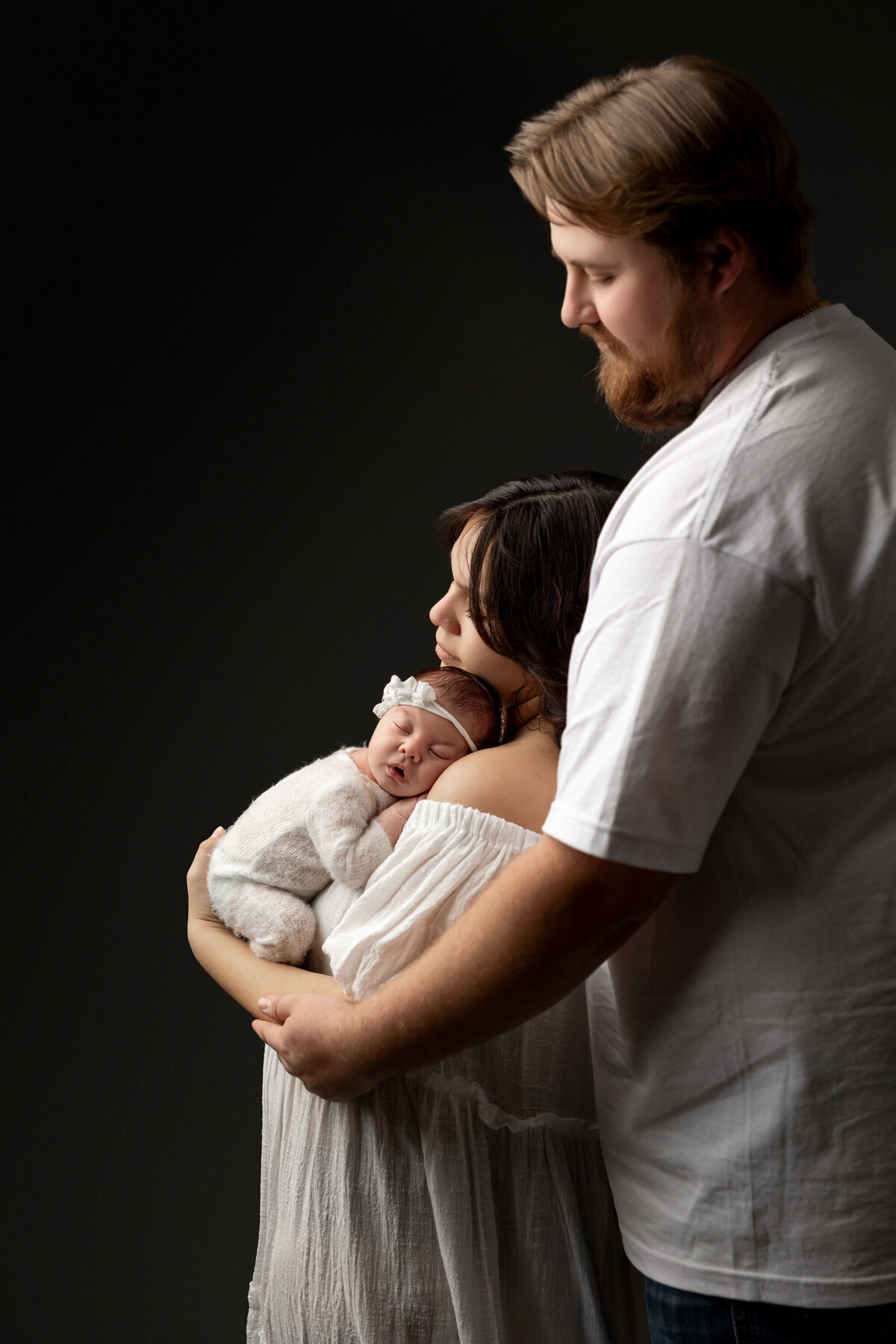 Parents holding their newborn baby girl close in a warm embrace, styled in white knits against a dark backdrop during their studio session. Captured by Inland Empire newborn photographer Kaitlyn Dawn Photography.