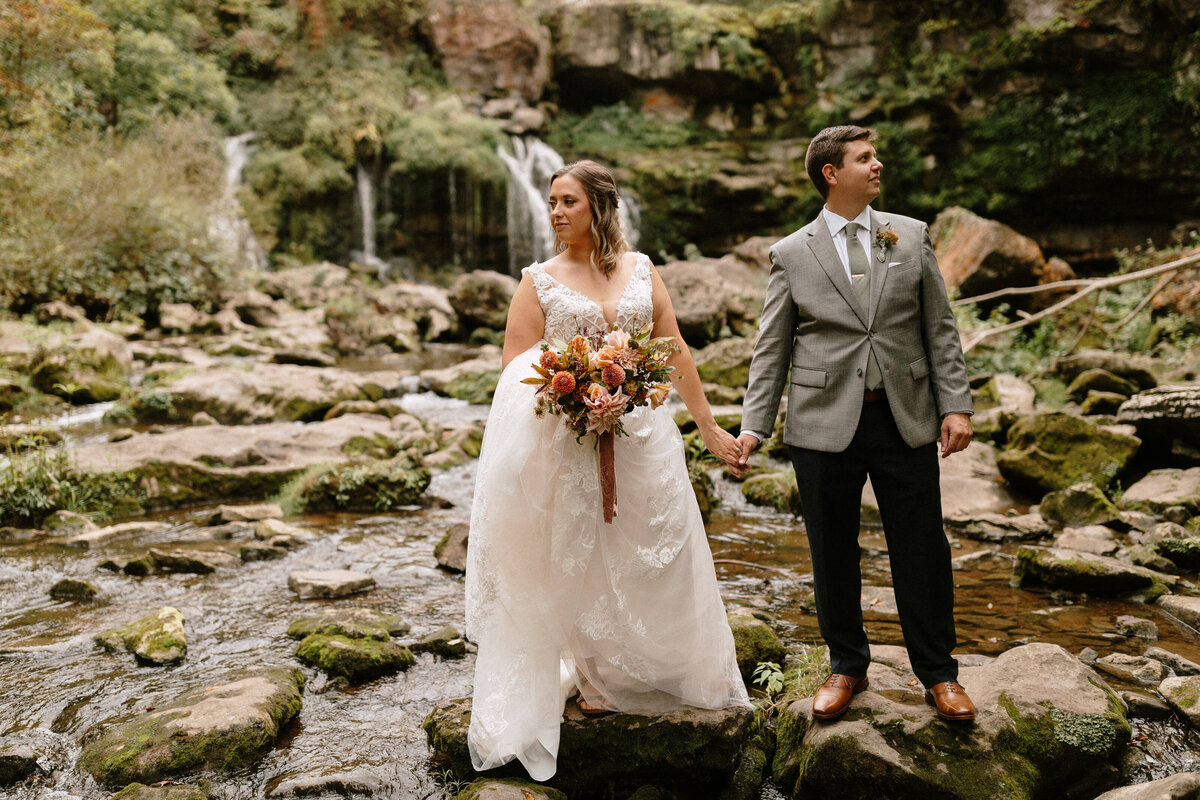 bride and groom stand hand in hand taking in the scenery in front of a waterfall