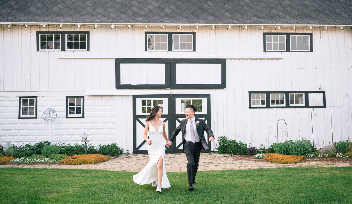 A Asian bride and groom run through the grass near a black and white barn