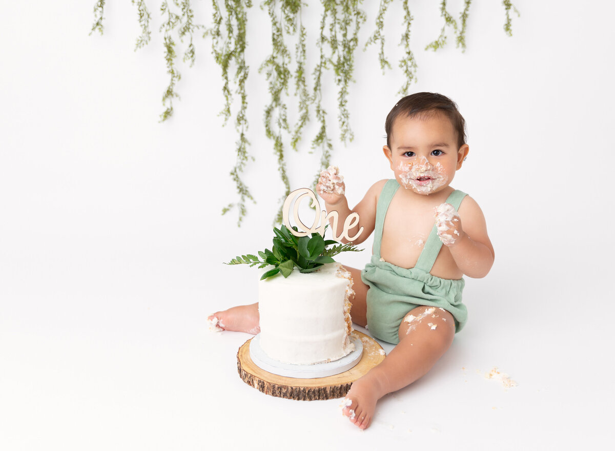 Baby boy in green shortalls sits with white cake topped with greenery between his legs. He has icing on his face and hands.