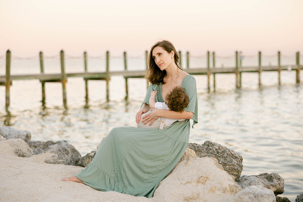 A mother in a flowing green dress sits on a rocky shore, breastfeeding her baby during a peaceful sunset in the Florida Keys. Captured by Claudia Amalia, Florida Keys family photographer.
