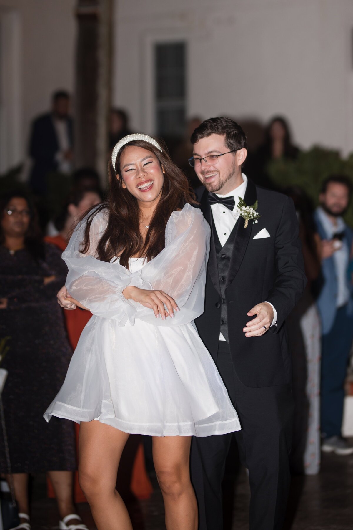 The bride and groom share a joyful laugh while dancing together during their wedding reception at Casa Marina Hotel in Jacksonville Beach, Florida. The bride wears a fun and flirty white mini dress with sheer sleeves and a pearl headband, while the groom is in a classic black tuxedo. Surrounded by friends and family, their happiness lights up the evening celebration. Captured by Heartful Films, a Jacksonville wedding photographer and videographer known for documenting genuine, candid moments full of emotion and personality.