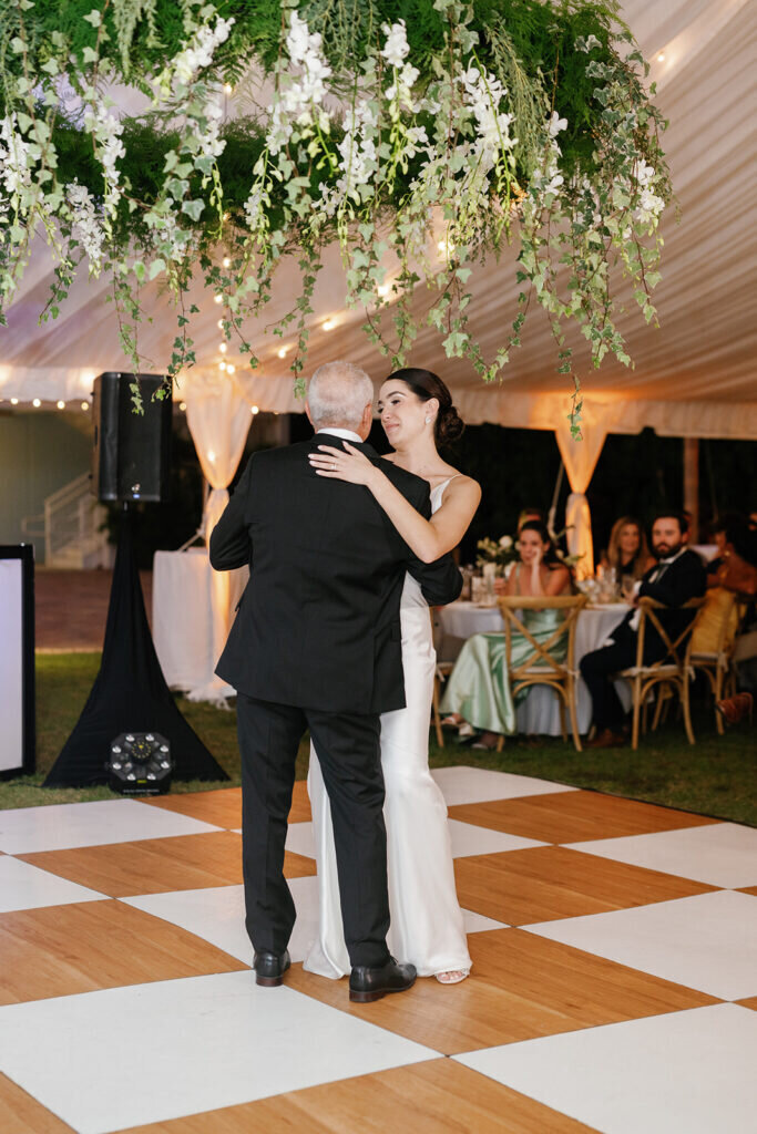 A touching wedding reception moment as the bride and her father embrace on the dance floor, surrounded by lush floral arrangements and warm ambient lighting. Destination wedding photography by Claudia Amalia Photography.