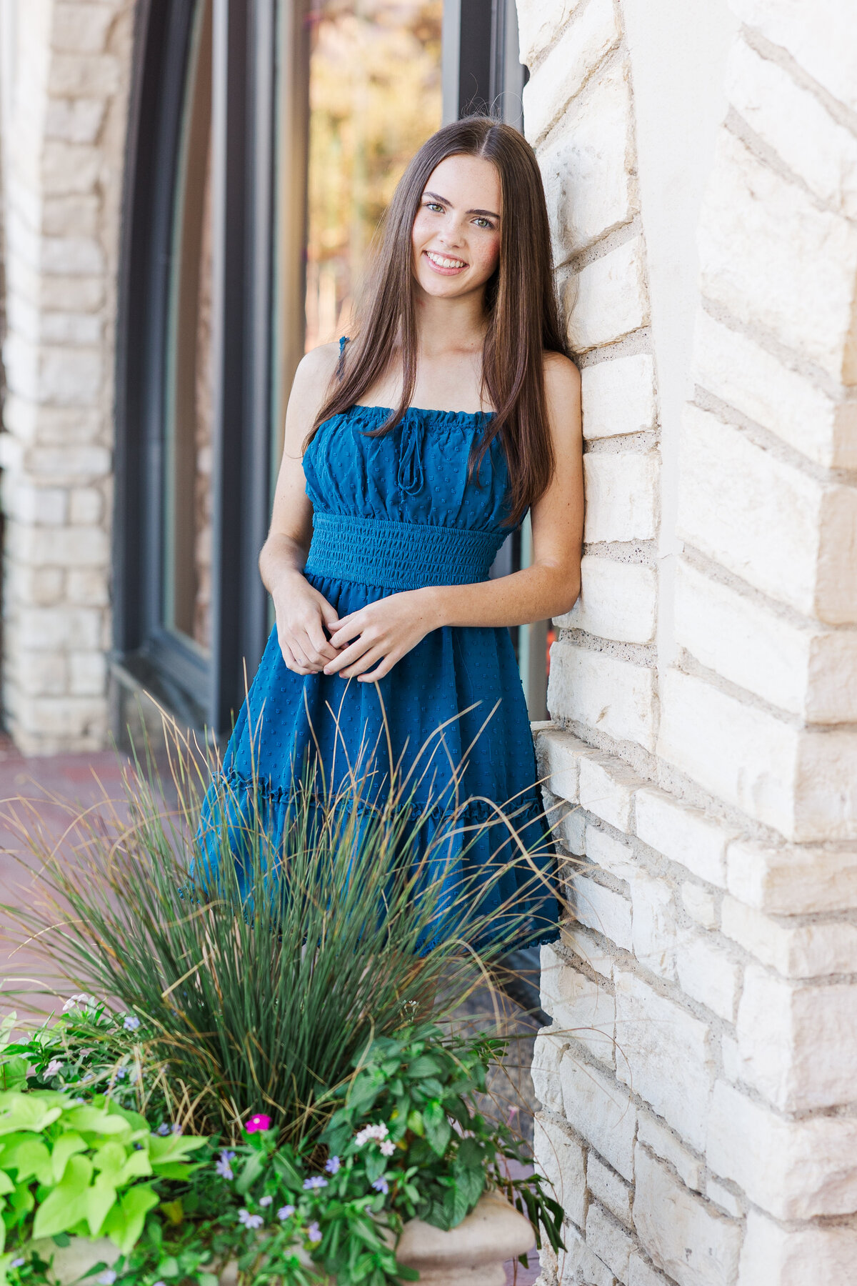 Senior portrait of a smiling girl in a teal blue sundress, standing beside a stone wall with green plants in the foreground. Captured in natural light in downtown Plano, Texas, with a relaxed and confident pose.