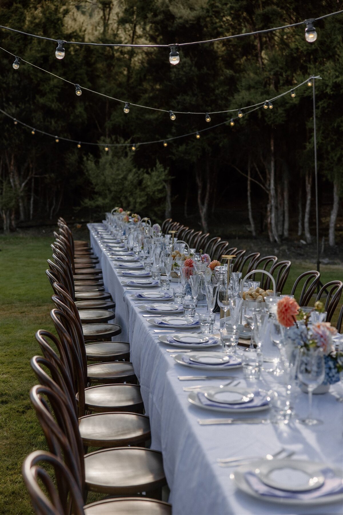 Elegant outdoor dining table with bentwood chairs, white linens, and crystal glassware set up for a Wanaka alfresco wedding reception.