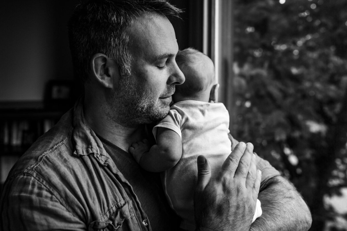 dad with eyes closed comforting newborn by window