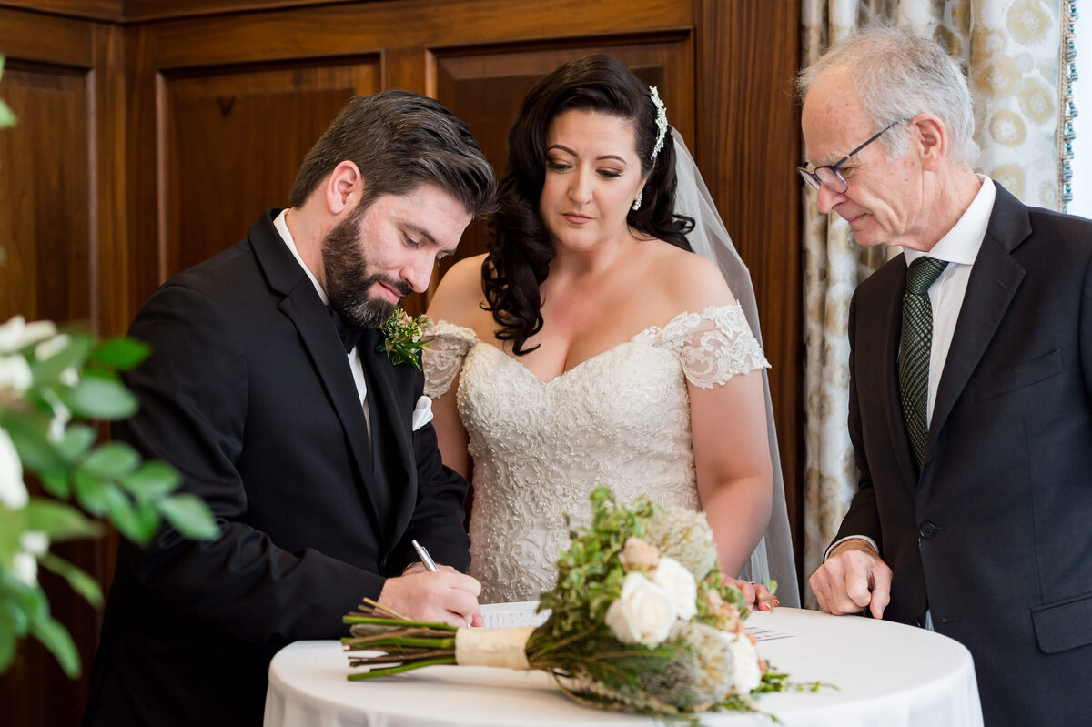 a groom in a black tuxedo signs his Ottawa wedding register while his bride and officiant look on.  Taken indoors at the Chateau Laurier hotel
