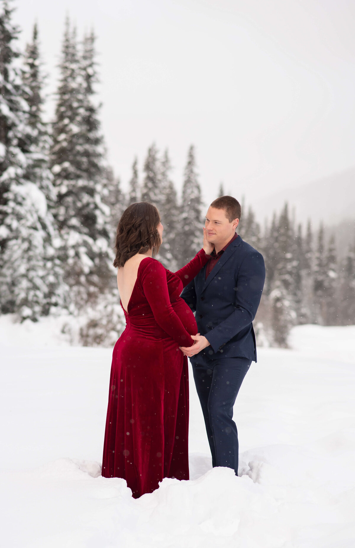 A winter backdrop sets the scene for this portrait of a husband and wife. She is pregnant and wearing a red dress. He is in a suit and they are looking at eachother holding hands. Taken by Brittany Danielle