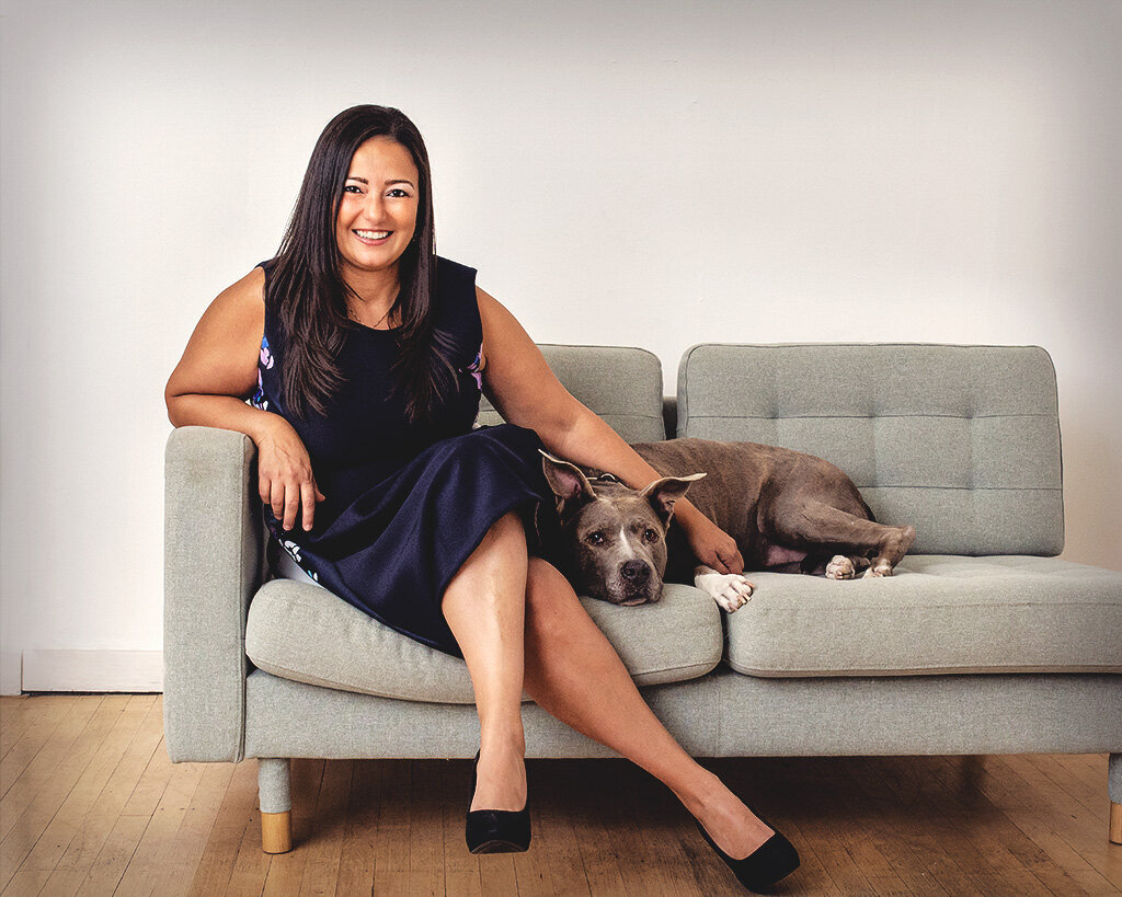 branding photo of a woman with dark hair in a blue dress sits on a grey coutch witht a grey dog