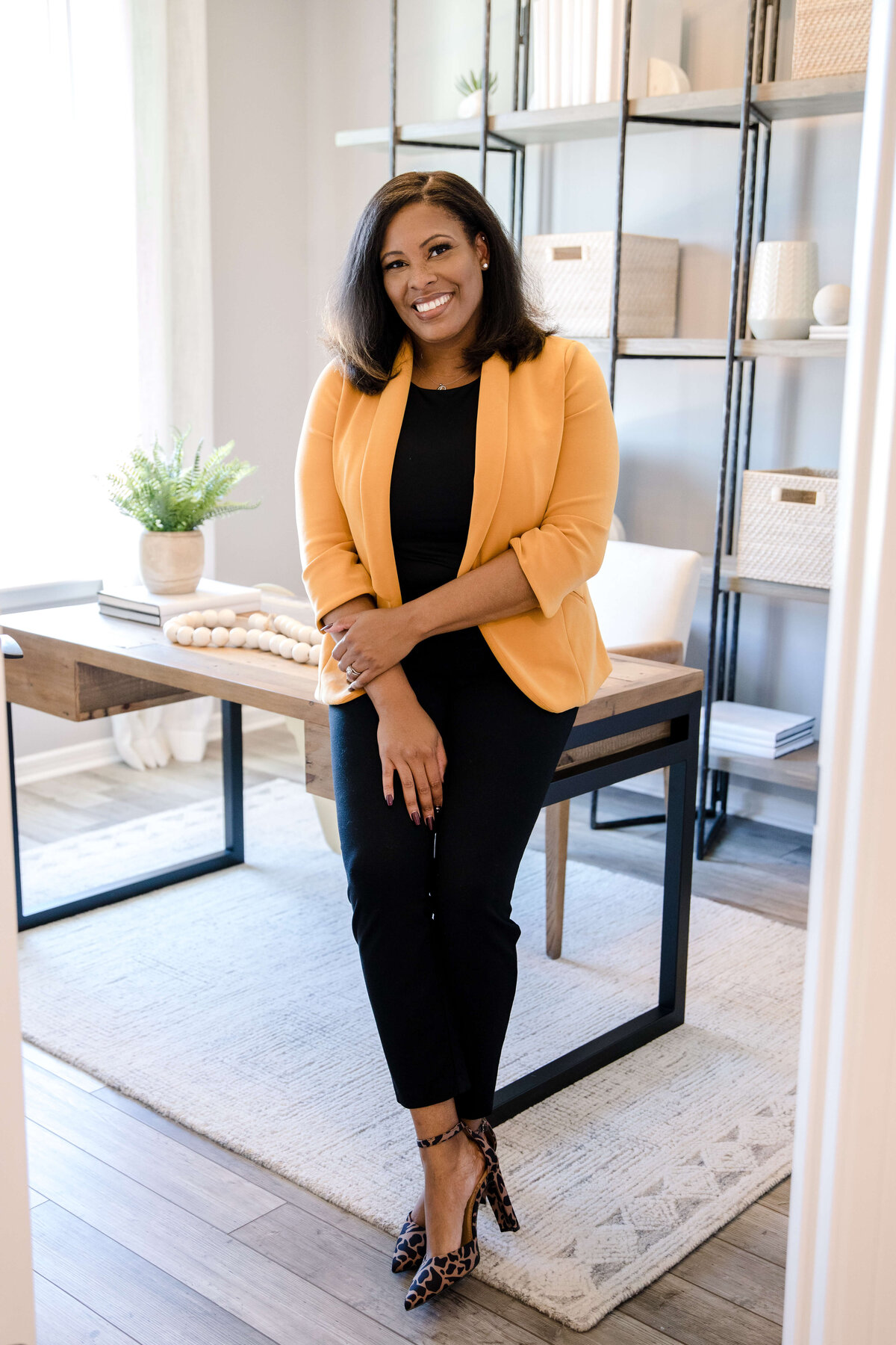 headshot portraits with woman leaning against a desk in a yellow blazer and a black jumper smiling with shelving behind her for brand photography