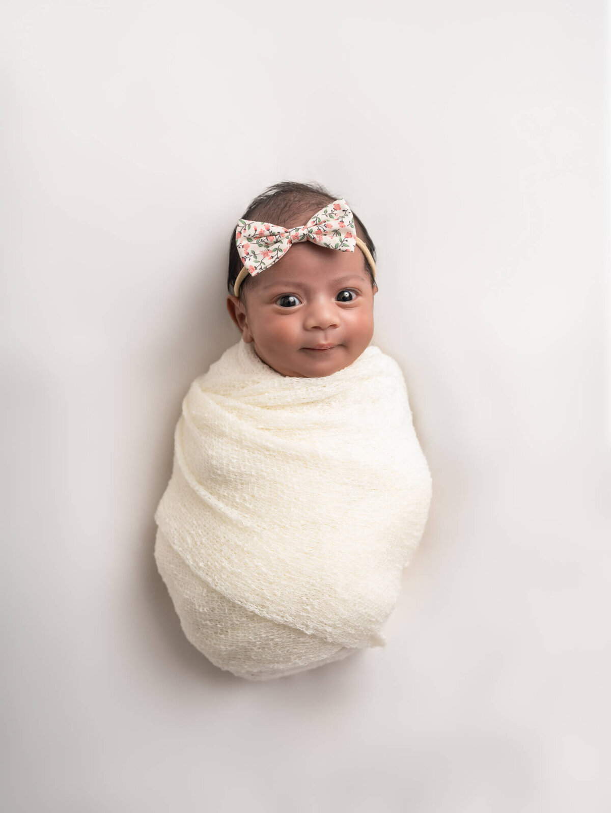 A newborn baby girl with a large floral bow and wrapped in a white blanket smiles with bright eyes