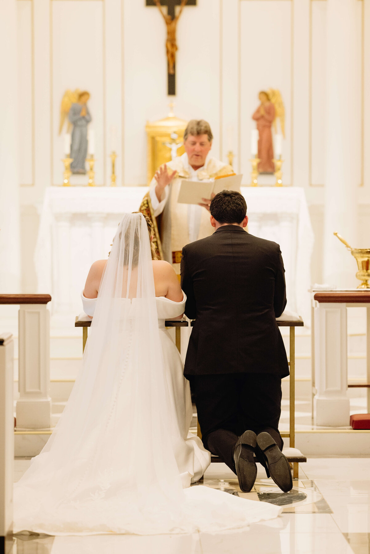 bride and groom kneeling at the alter during their wedding ceremony in a catholic mass with the priest blessing them captured by Virginia wedding photographer