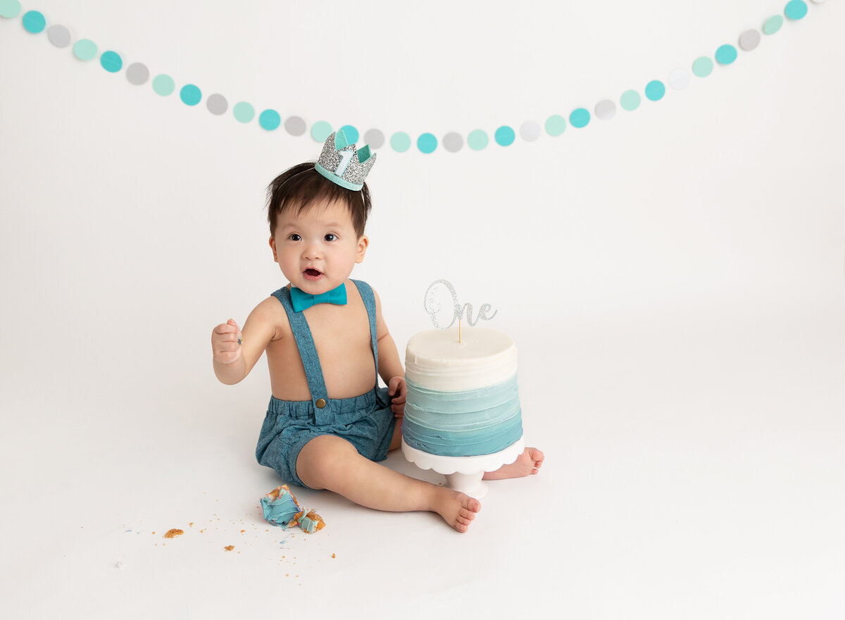 Baby boy in denim shorts with suspenders sits with blue ombre cake between his legs.