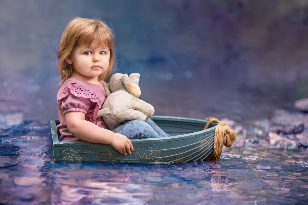 Whimsical portrait of a toddler sitting in a small boat surrounded by dreamy water reflections, holding a stuffed animal. Captured in Overland Park, Kansas, this setup is perfect for families seeking imaginative and themed child photography.