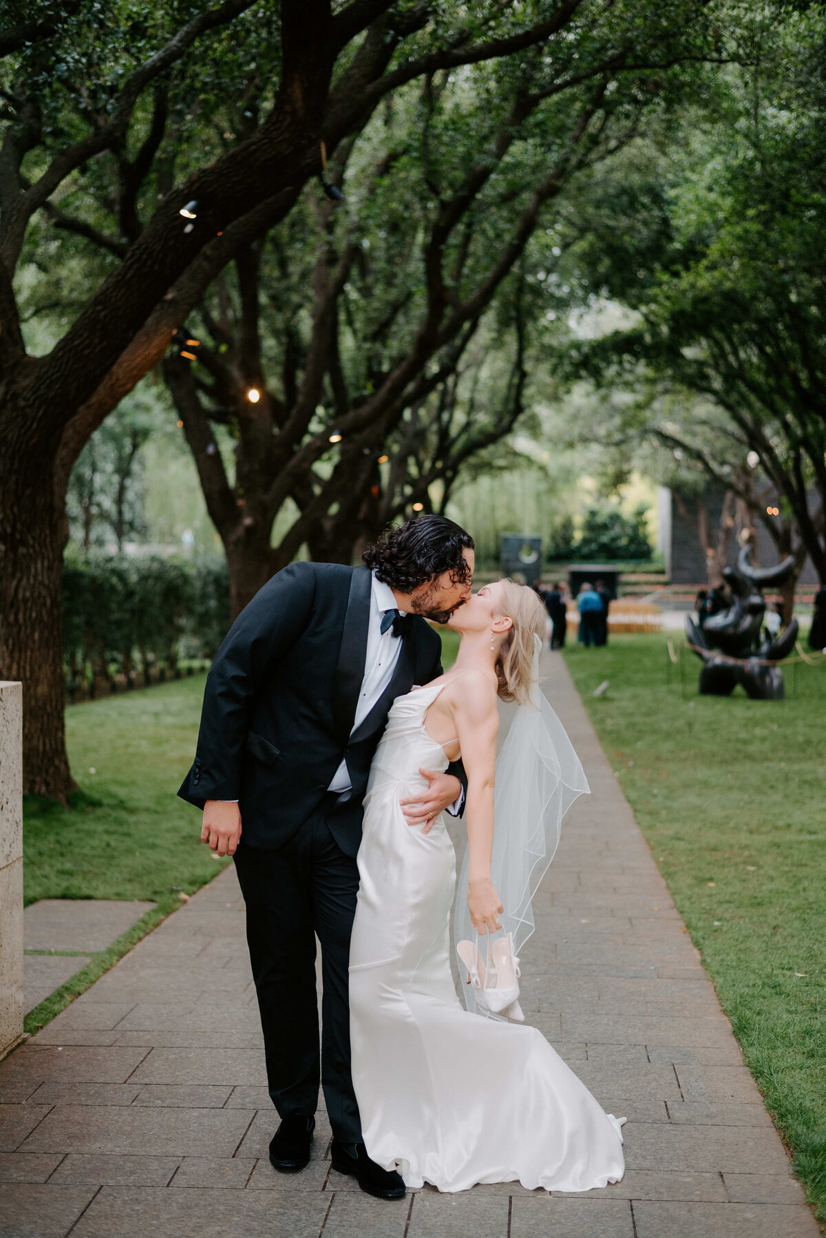 Bride and groom sharing a romantic kiss in a tree-lined garden pathway, with the groom dipping the bride in her satin wedding dress and veil