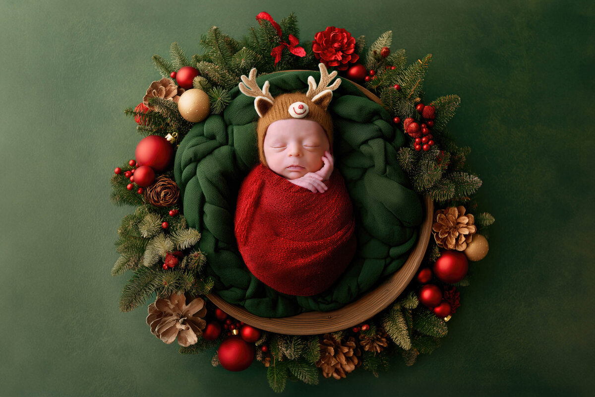 newborn boy wrapped in red wearing a reindeer hat in a wreath background for his Christmas newborn photography session.