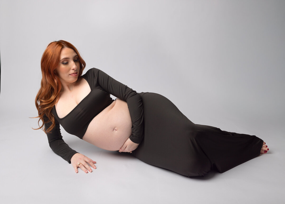 Pregnant women in a from fitting black dress holds her baby bump while laying on her side during a maternity session in Waldwick, NJ
