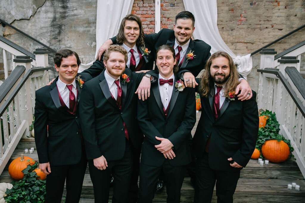Six men dressed in black suits with maroon vests and ties pose together, smiling, on a wooden staircase decorated with pumpkins, greenery, and white drapery. Exposed brick wall in the background.